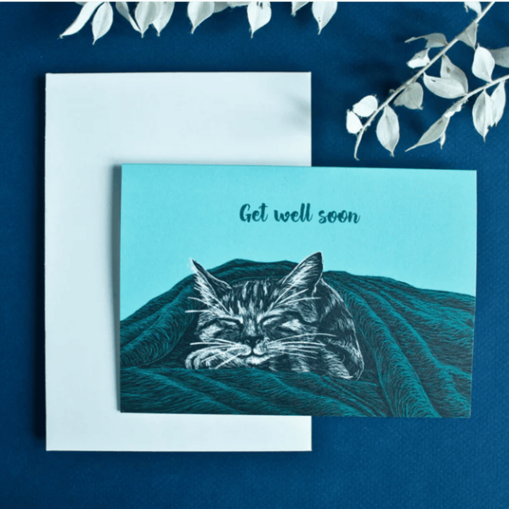 Illustrierte Postkarte in türkis mit einer Katze, die zufrieden unter einer Decke schläft. Aufschrift: "Get well soon"