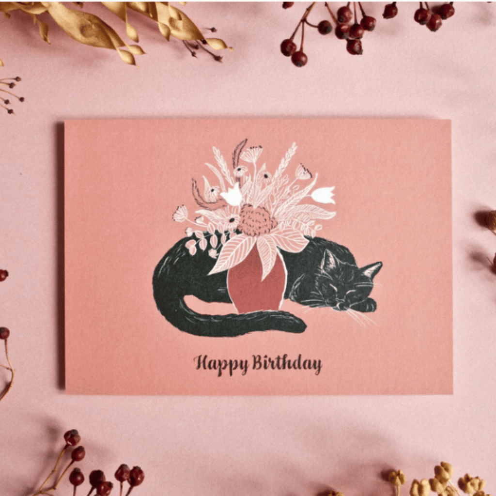 Illustrierte Postkarte mit einer schwarzen Katze, die um einen Blumenstrauß in einer Vase liegt. Aufschrift: "Happy Birthday"