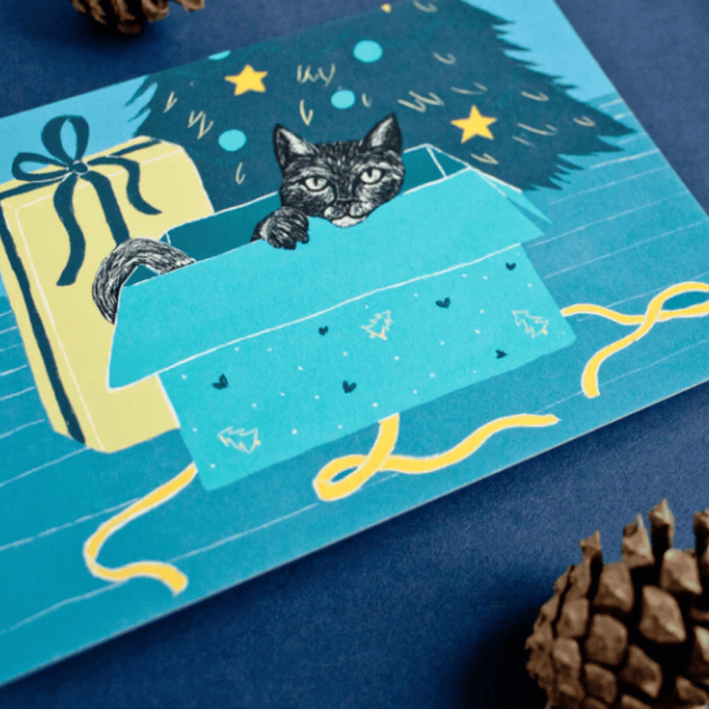 Illustrierte Postkarte mit einer schwarzen Katze in einem ausgepackten Geschenk vor einem Weihnachtsbaum