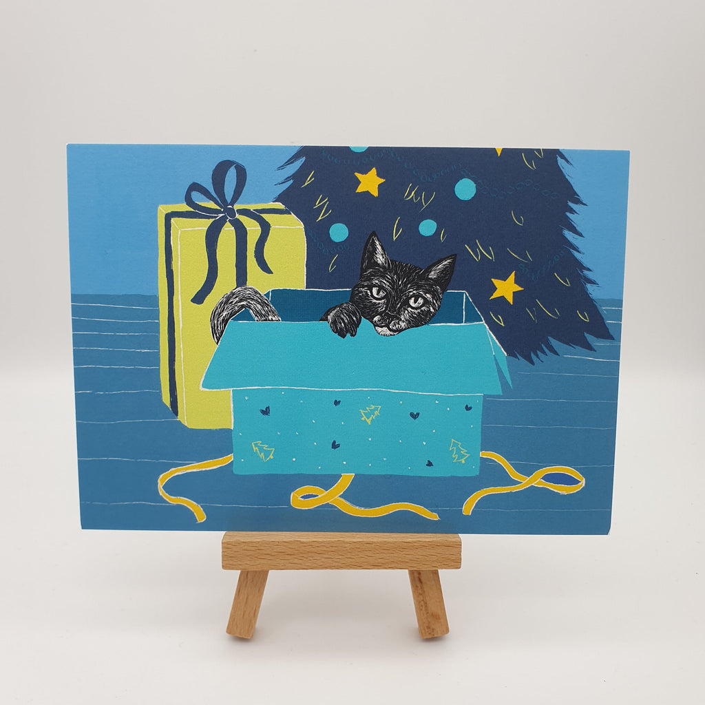 Illustrierte Postkarte mit einer schwarzen Katze in einem ausgepackten Geschenk vor einem Weihnachtsbaum