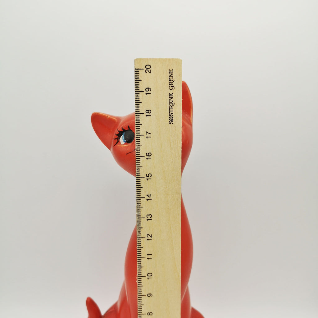 Rote Figur aus Keramik in Form einer schlanken sitzenden Katze hinter einem Lineal