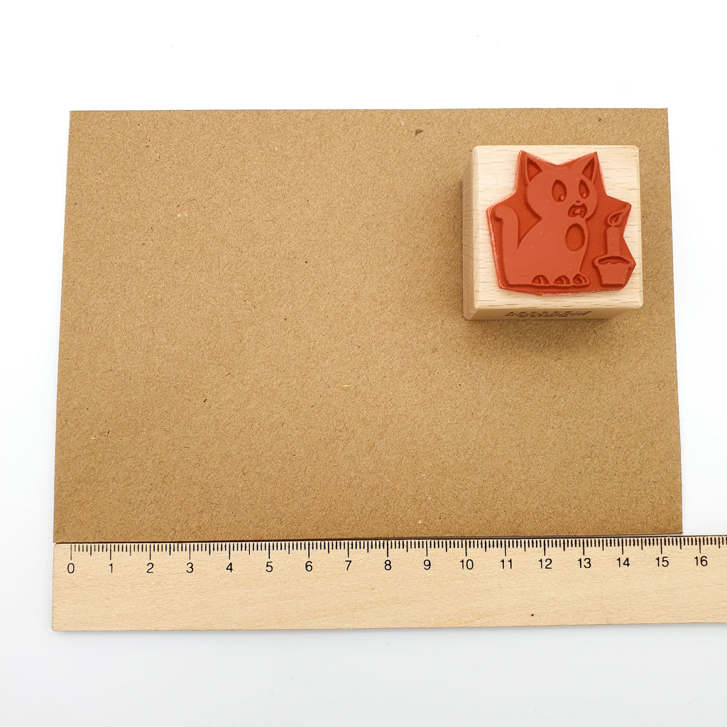 Stempel aus rotem Gummi mit dem Motiv einer Katze, die eine Kerze ausbläst auf einem braunen Briefumschlag mit Lineal