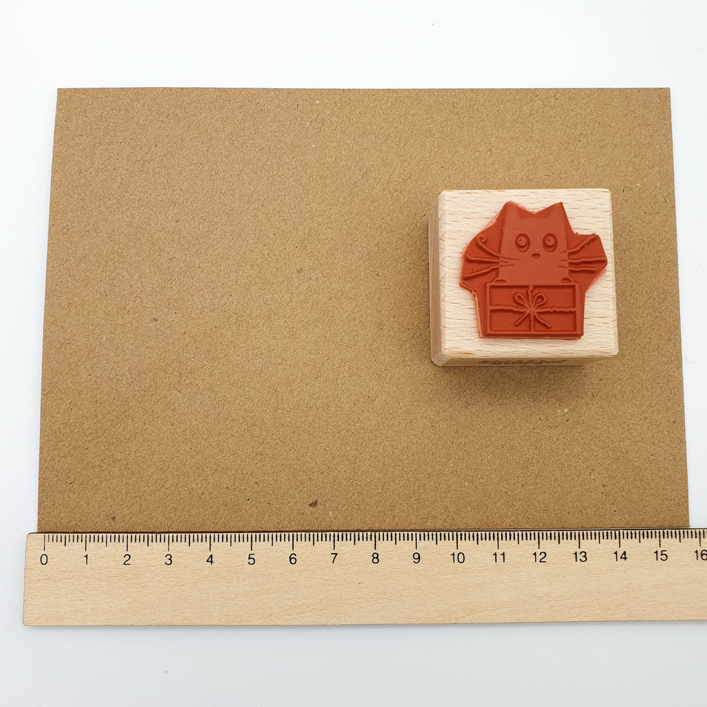 Stempel aus rotem Gummi mit dem Motiv einer Katze, die in einem Geschenk sitzt auf einem braunen Briefumschlag mit Lineal