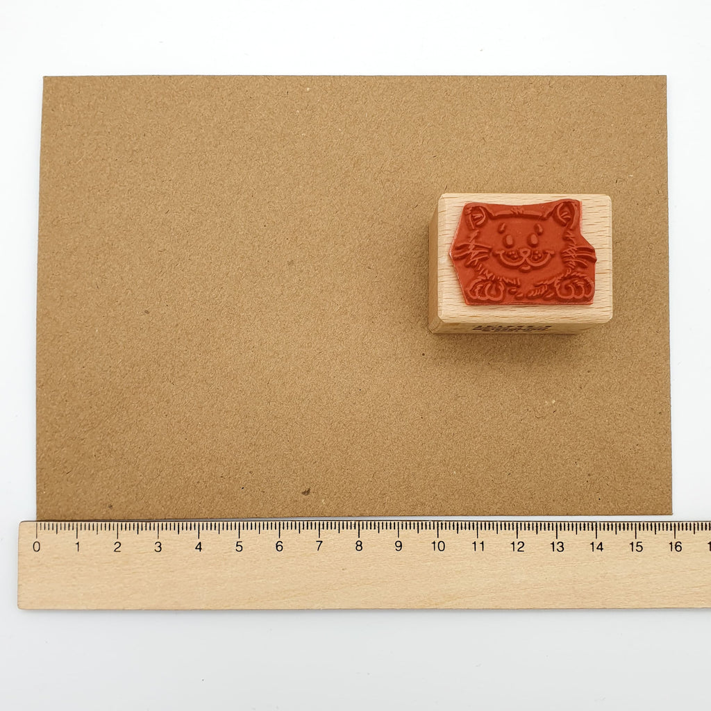 Stempel aus rotem Gummi mit dem Motiv einer lächelnden Katze auf einem braunen Briefumschlag mit Lineal